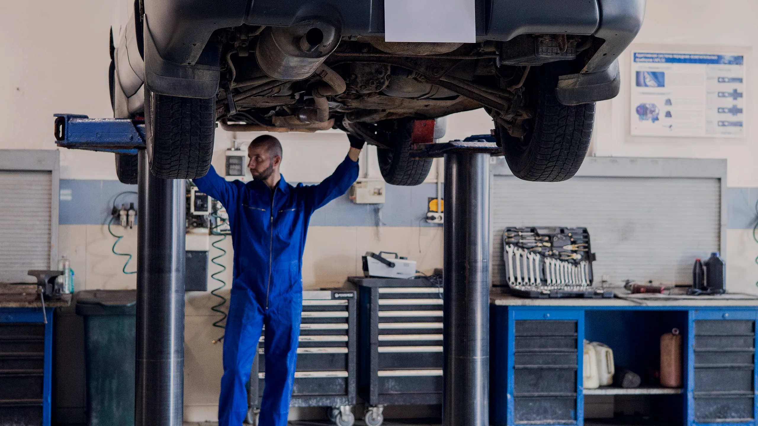 A mechanic in blue overalls standing beneath a car raised on a two-post lift, one hand against the underbody, in a bright workshop with tool chests along the wall.