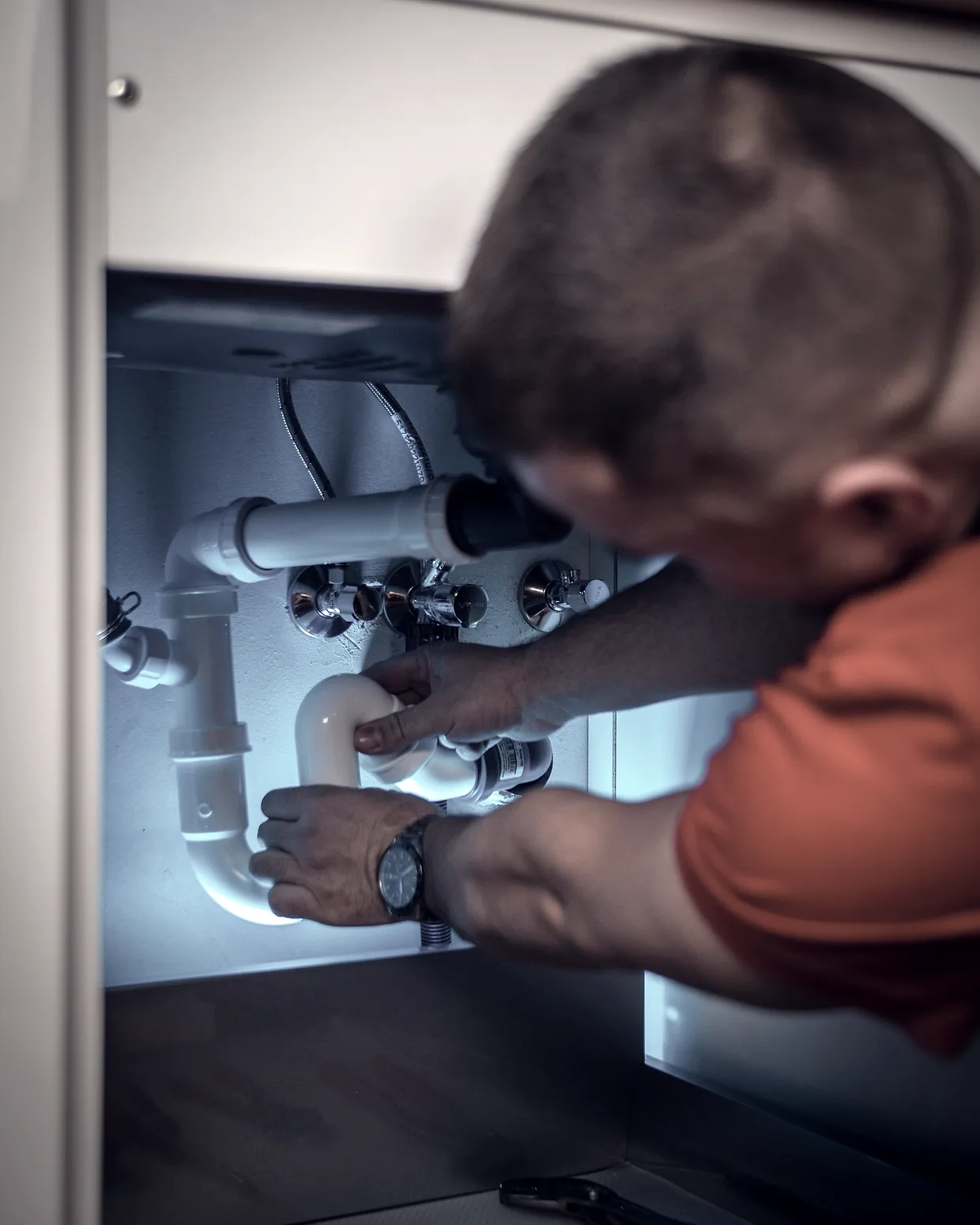A plumber reaching into the cabinet under a kitchen sink, both hands on the drain trap.