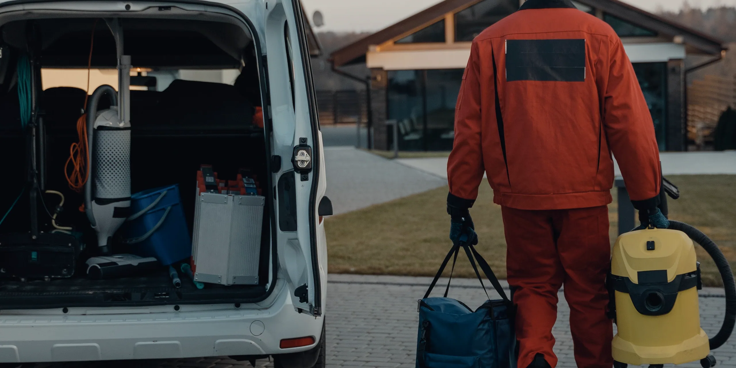 A cleaner carrying equipment from the back of a van toward a house.