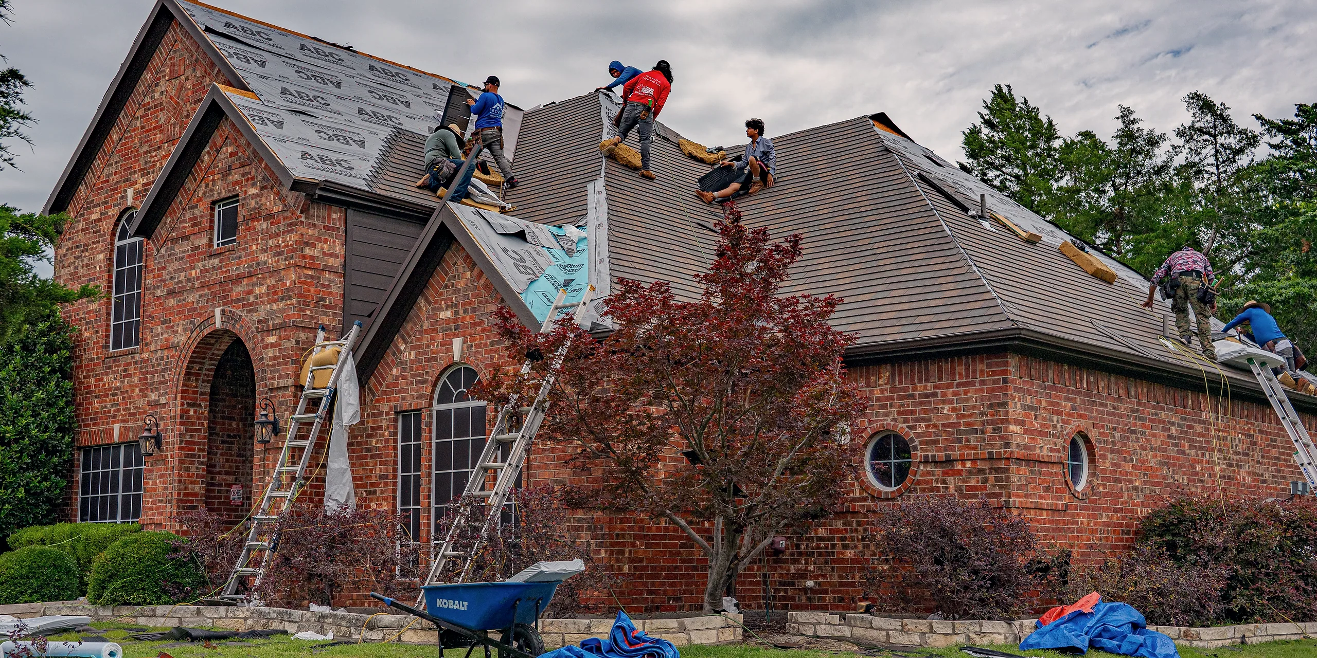 A roofing crew stripping and replacing the roof of a brick house, ladders up and tarps down.